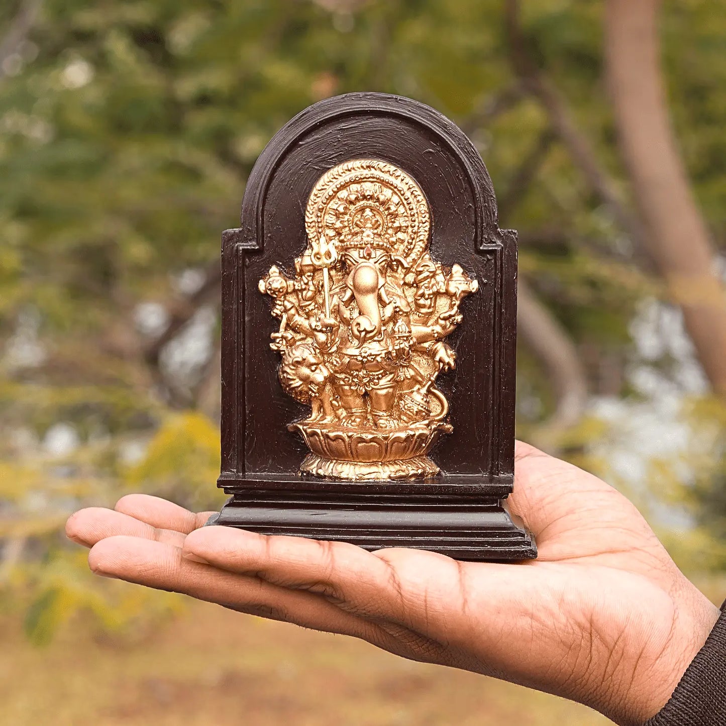 Hand holding a small golden deity statue on a dark wooden base against a blurred natural background