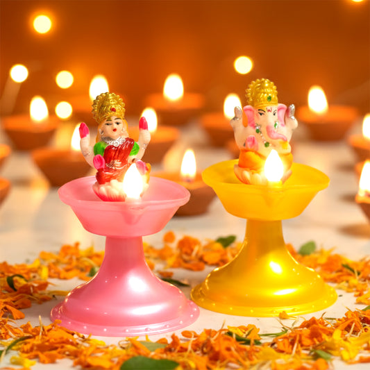 Two small figurines on pink and yellow stands with candles, surrounded by flowers and blurred lights in the background. Water Sensor Laxmi ganesh Diya.