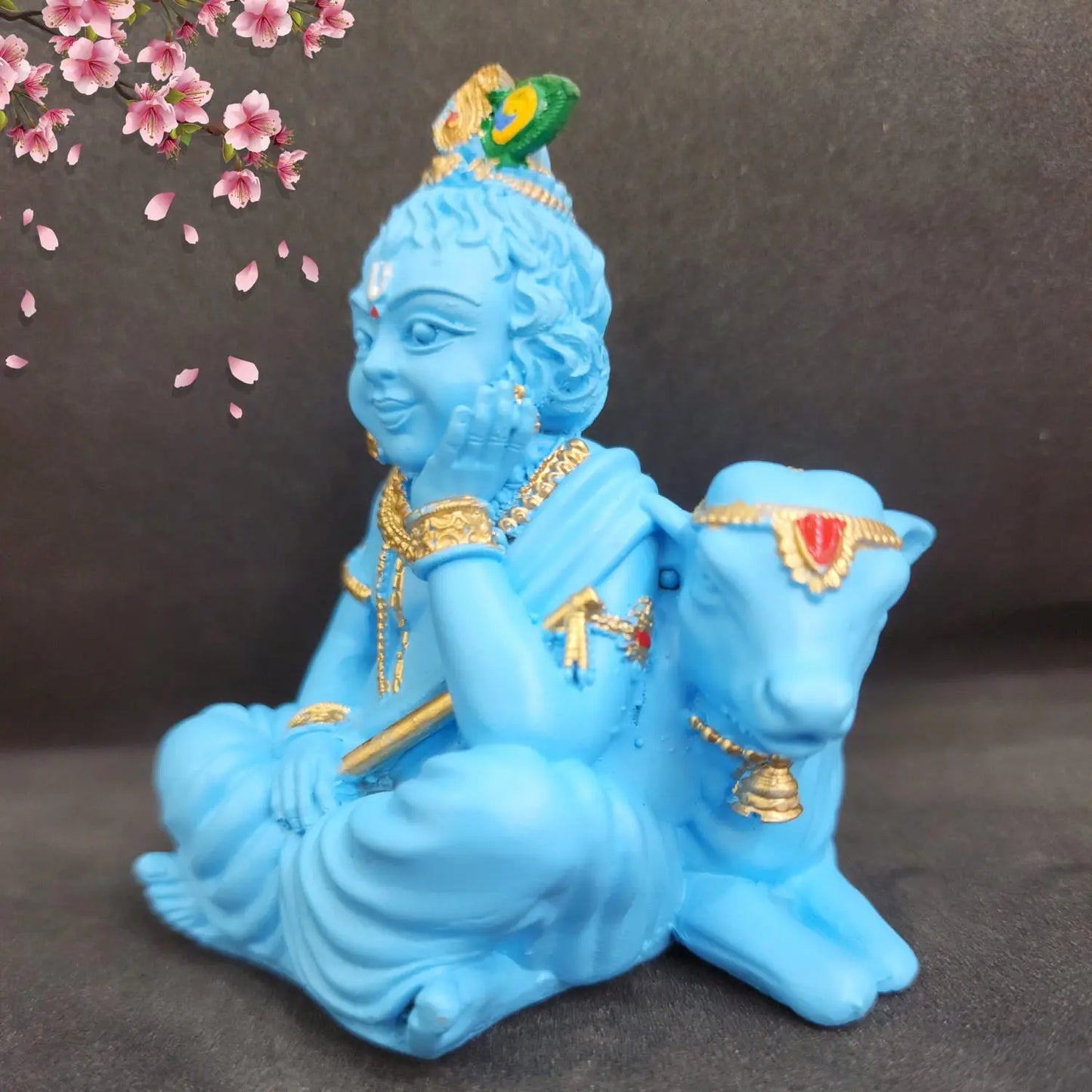 Blue statue of a seated figure with a cow, decorated with gold and red details, against a dark background with cherry blossoms.