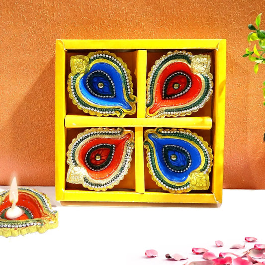 Colorful Diwali decorative items in a yellow box with a lit candle and rose petals on a white surface.