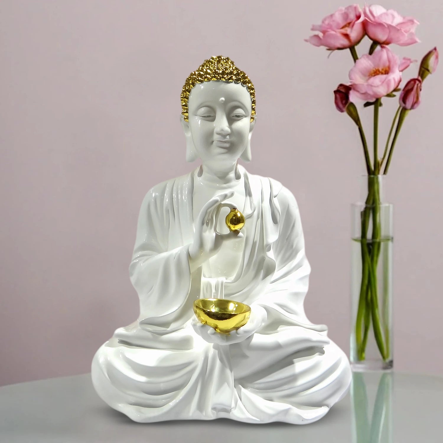 White Buddha statue with gold accents holding a bowl, next to a vase of pink flowers on a light pink background