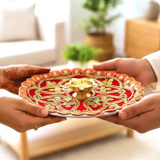 Two hands holding a decorative red and gold plate with a small lamp inside, set against a blurred indoor background.