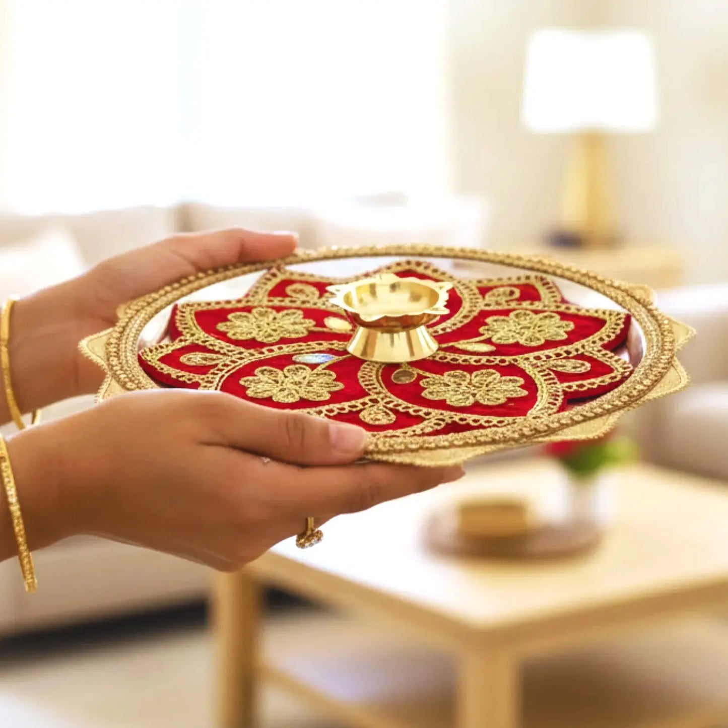 Two hands holding a decorative red and gold plate with a small lamp inside, set against a blurred indoor background.