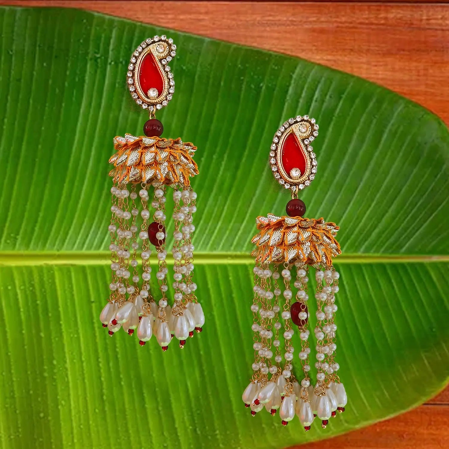 Pair of ornate earrings with red stones and pearl-like beads on a green leaf background