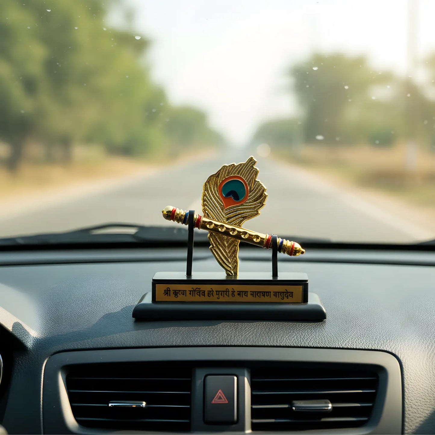 Decorative peacock feather ornament on a car dashboard with a blurred road and trees in the background.