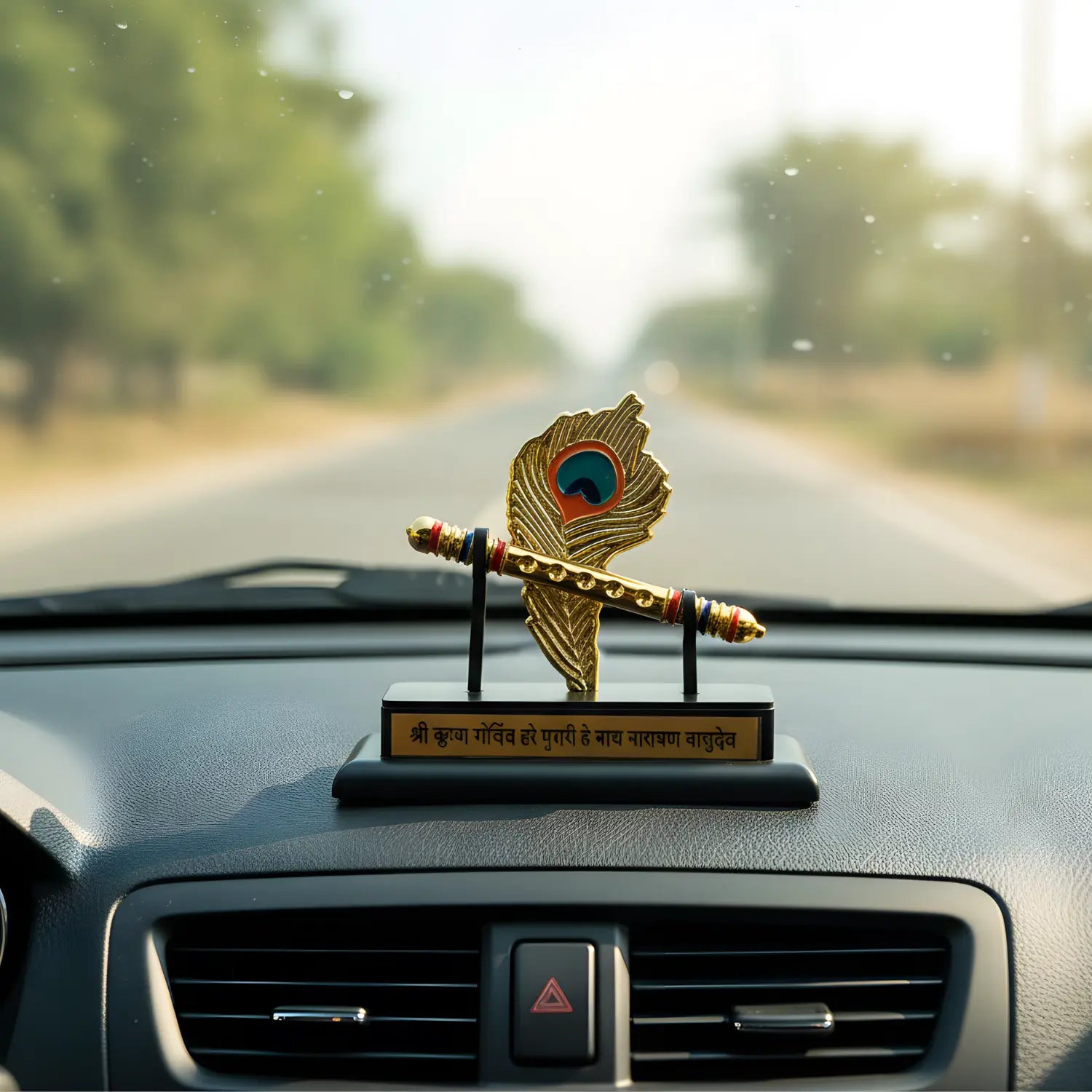 Decorative peacock feather ornament on a car dashboard with a blurred road and trees in the background.