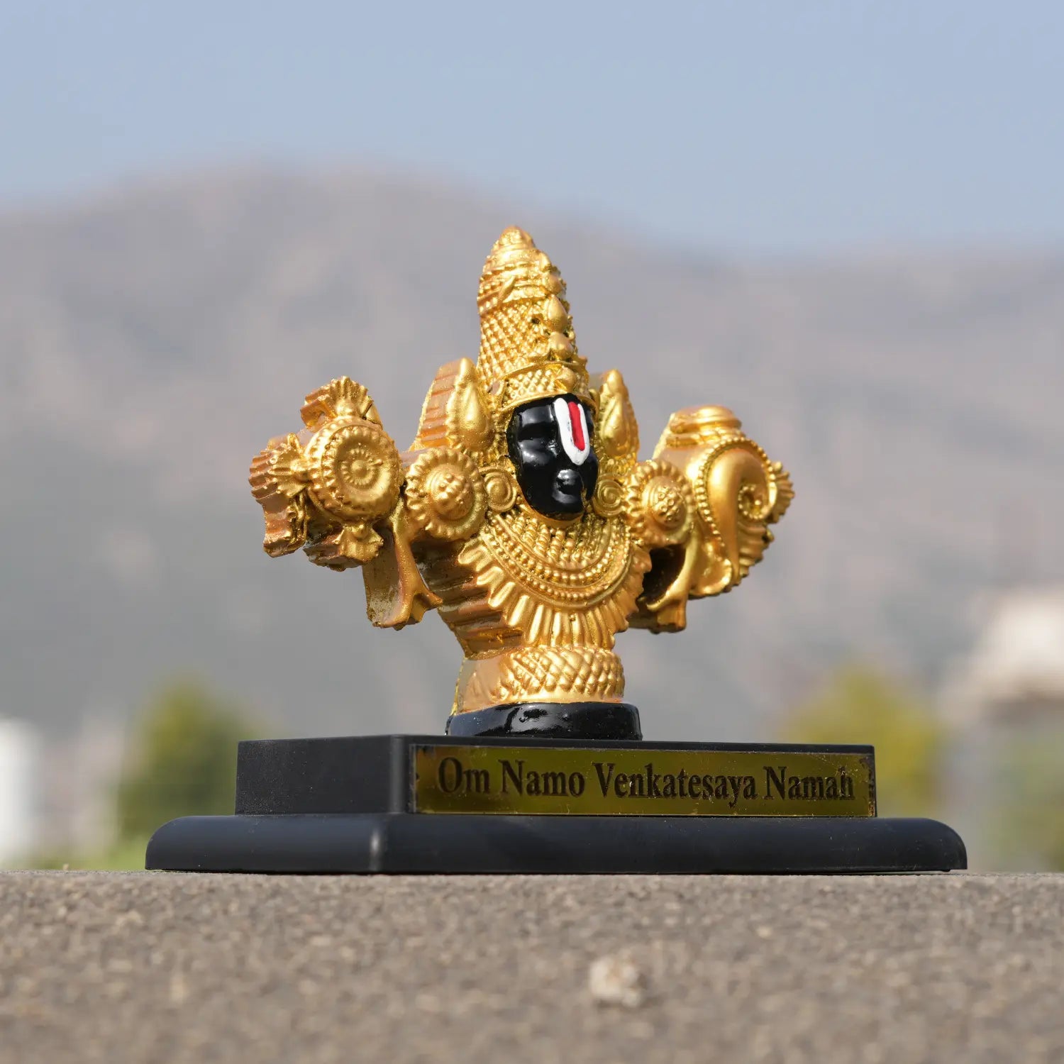 Gold-colored deity statue with 'Om Namo Venkatesaya Nama' text on a blurred natural background