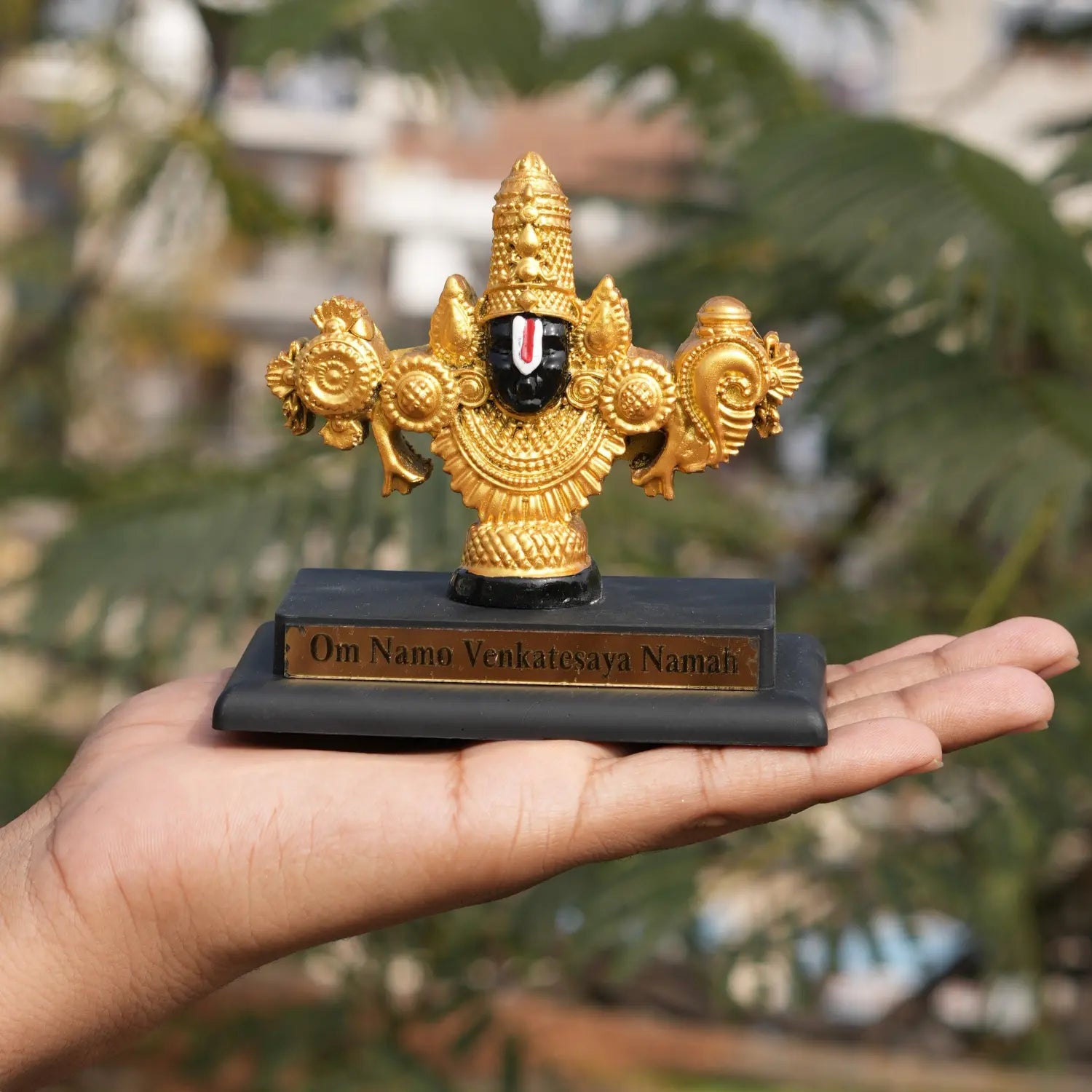 Decorative statue on a black base held in a hand with green foliage in the background