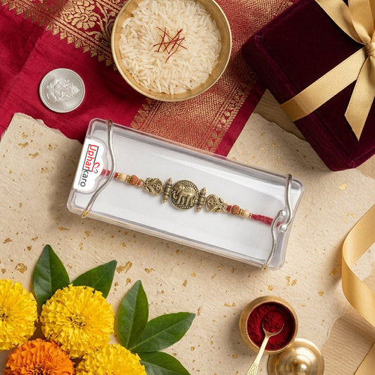 A traditional Indian Rakhi bracelet in a clear plastic packaging box, displayed on a decorated flat-lay background with a red gift box, rice, marigold flowers, and a coin.