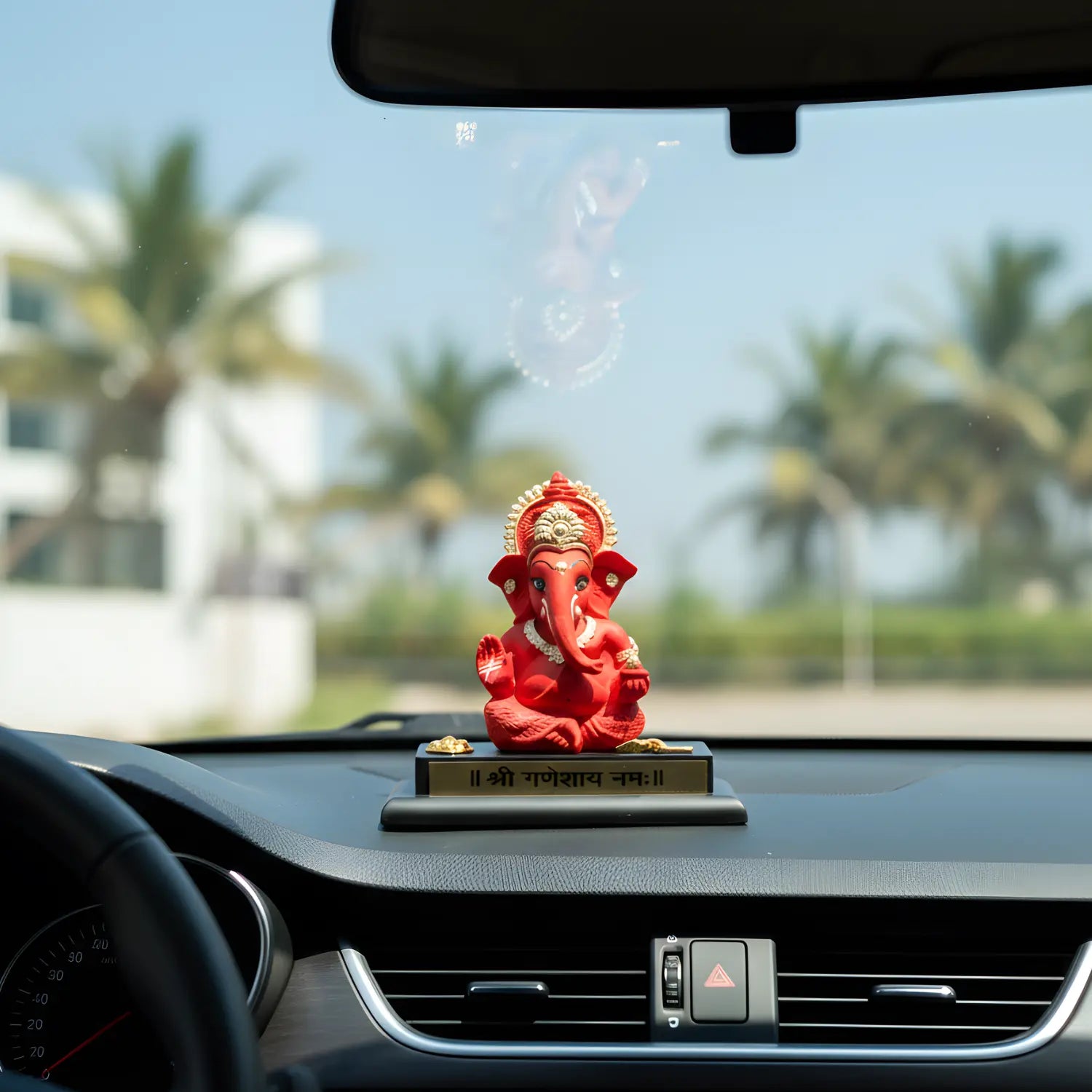 Red deity statue on a car dashboard with palm trees in the background