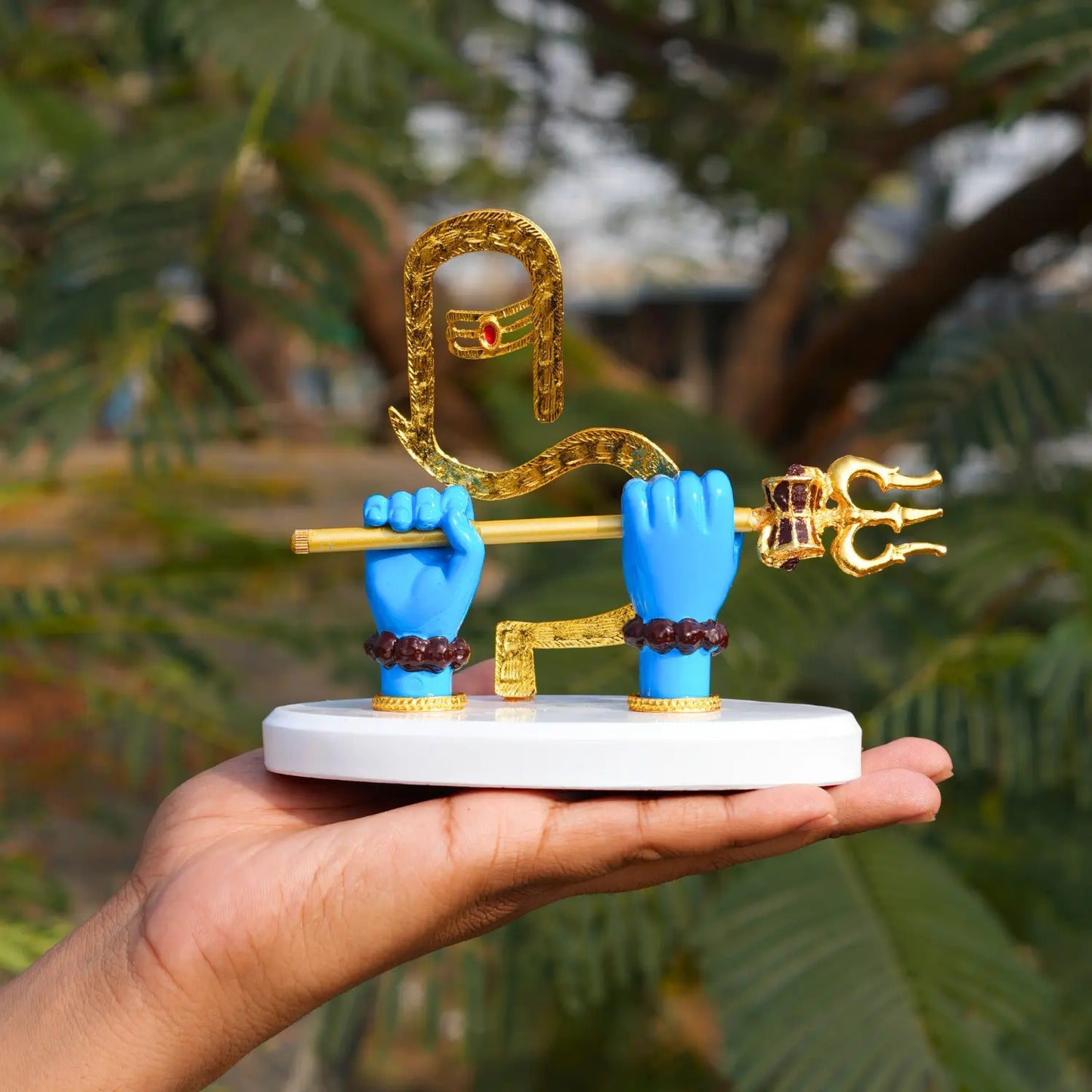 Small statue with blue hands holding a golden weapon on a white base, held by a person against a blurred green background.