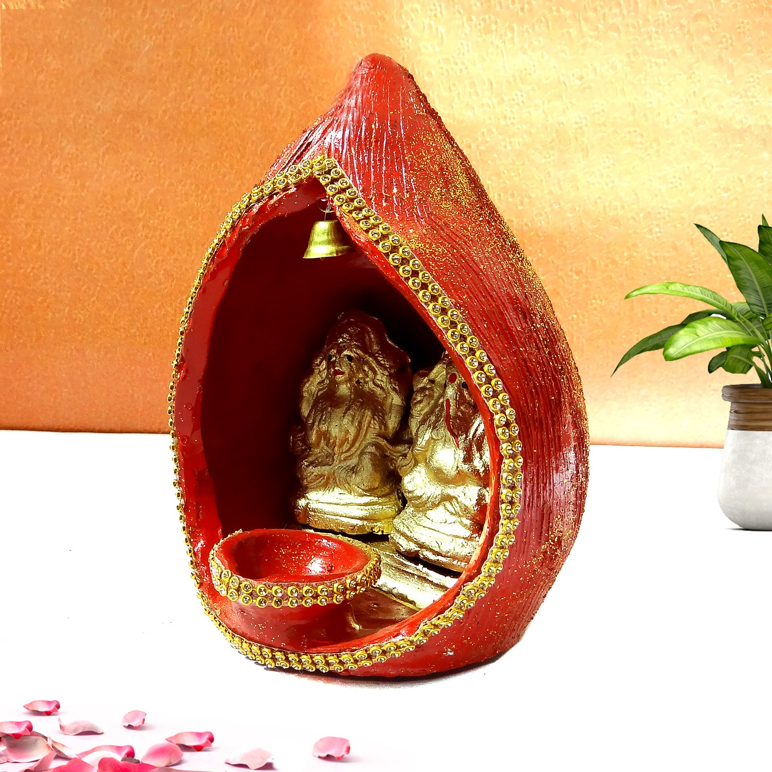 Decorative red and gold shrine with a small statue inside, placed on a white surface with a plant in the background.