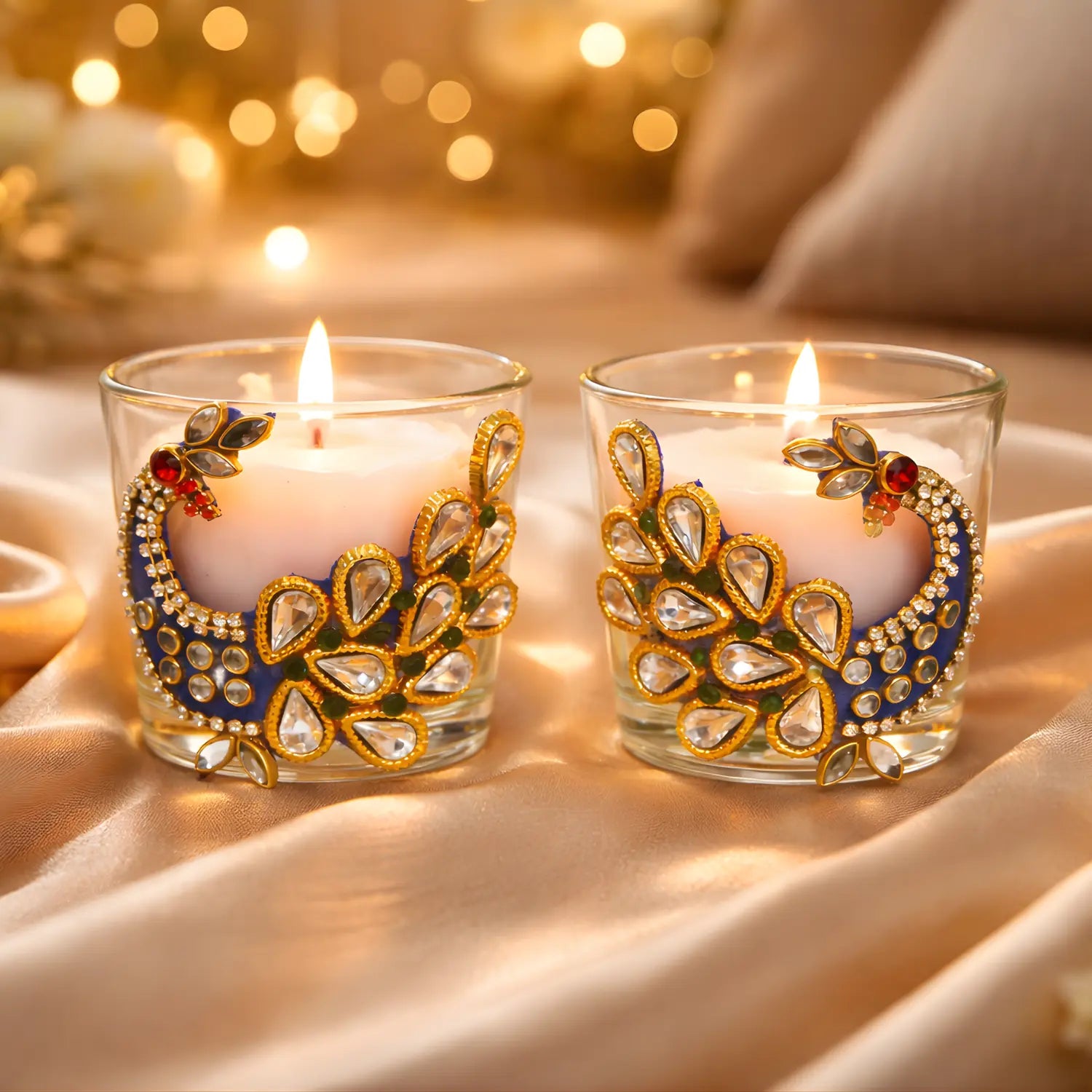 Two decorative candles in glass holders with peacock feather design on a soft fabric background.