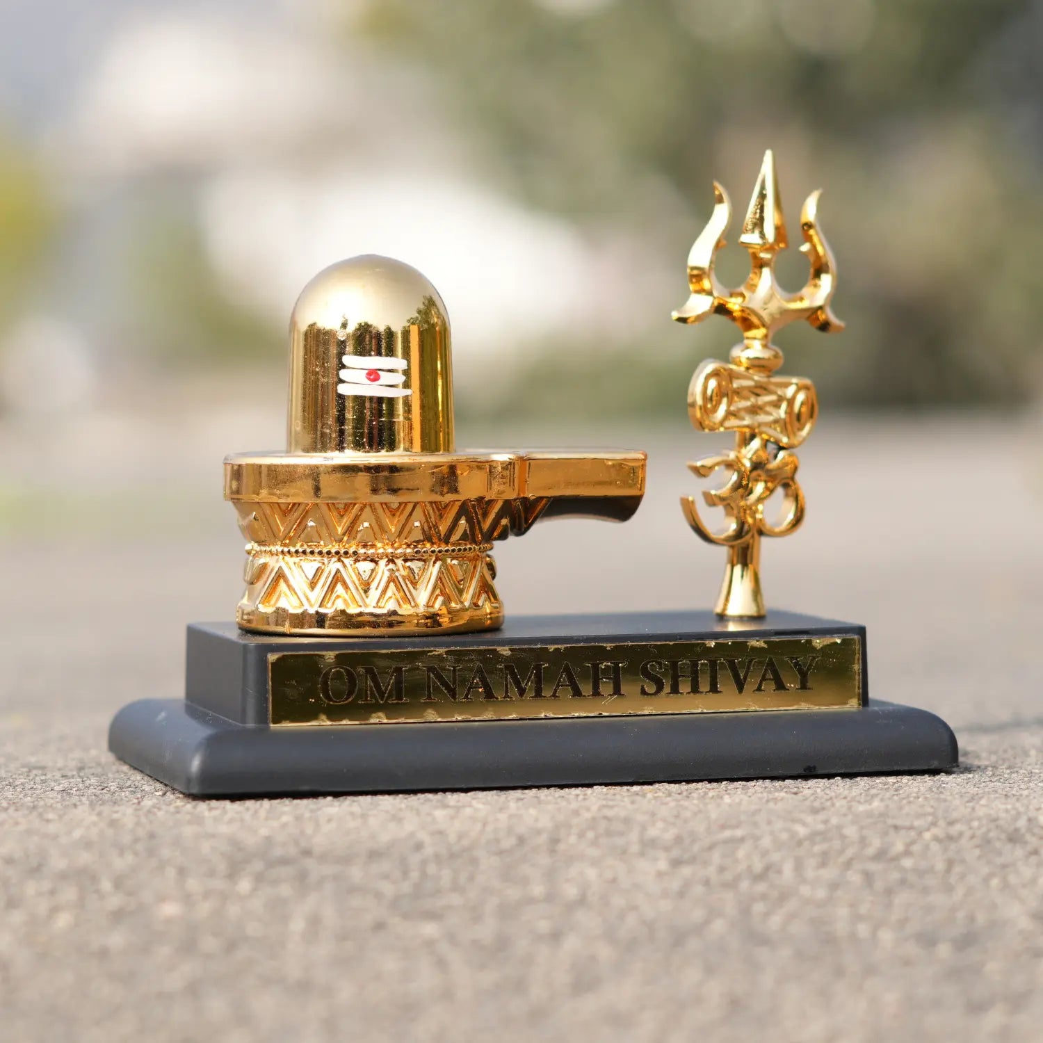 Gold Shiva Lingam and Trishul on a black base with 'OM NAMAH SHIVAY' inscription, outdoor setting.