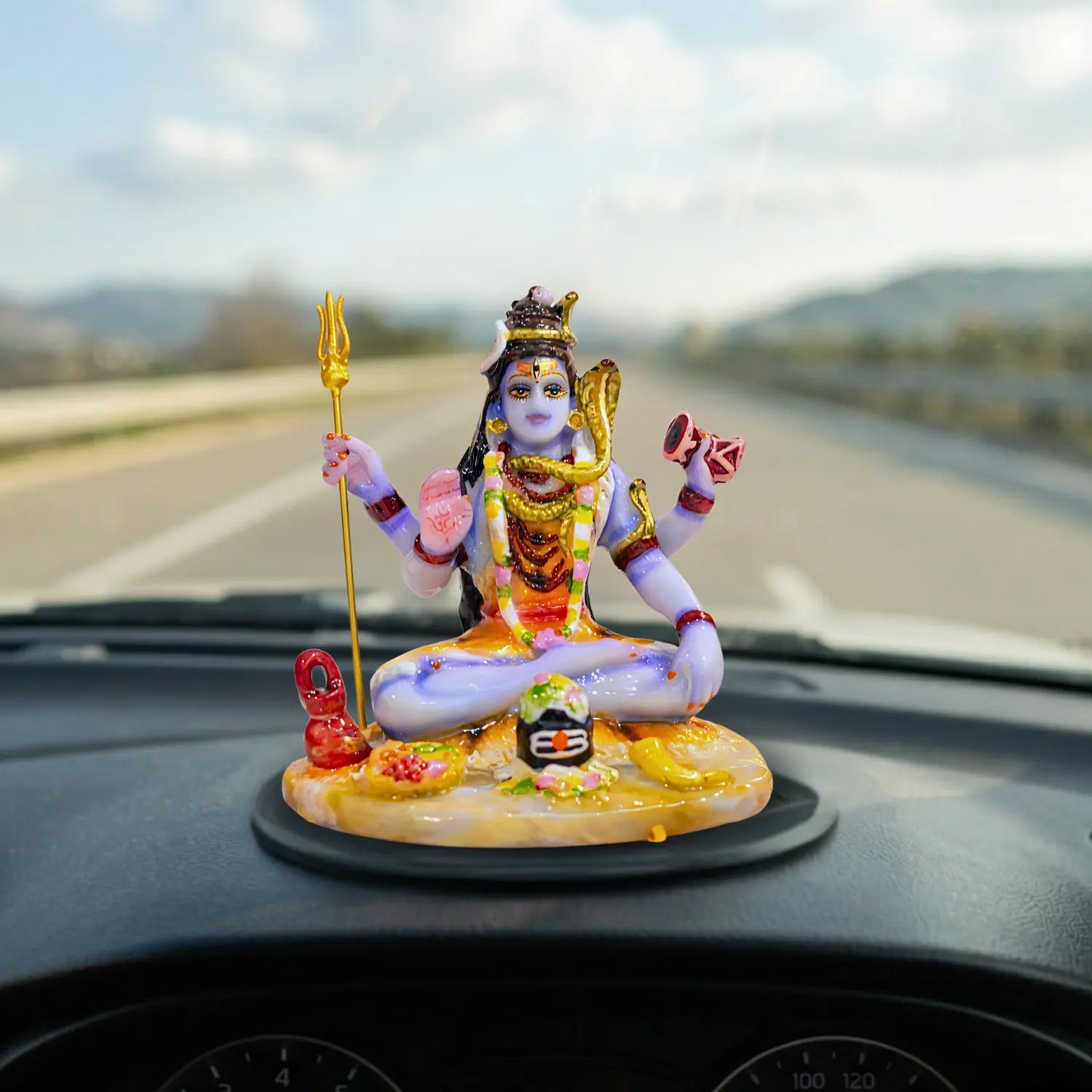 Decorative figurine of MEHADEV on a car dashboard with a blurred road background