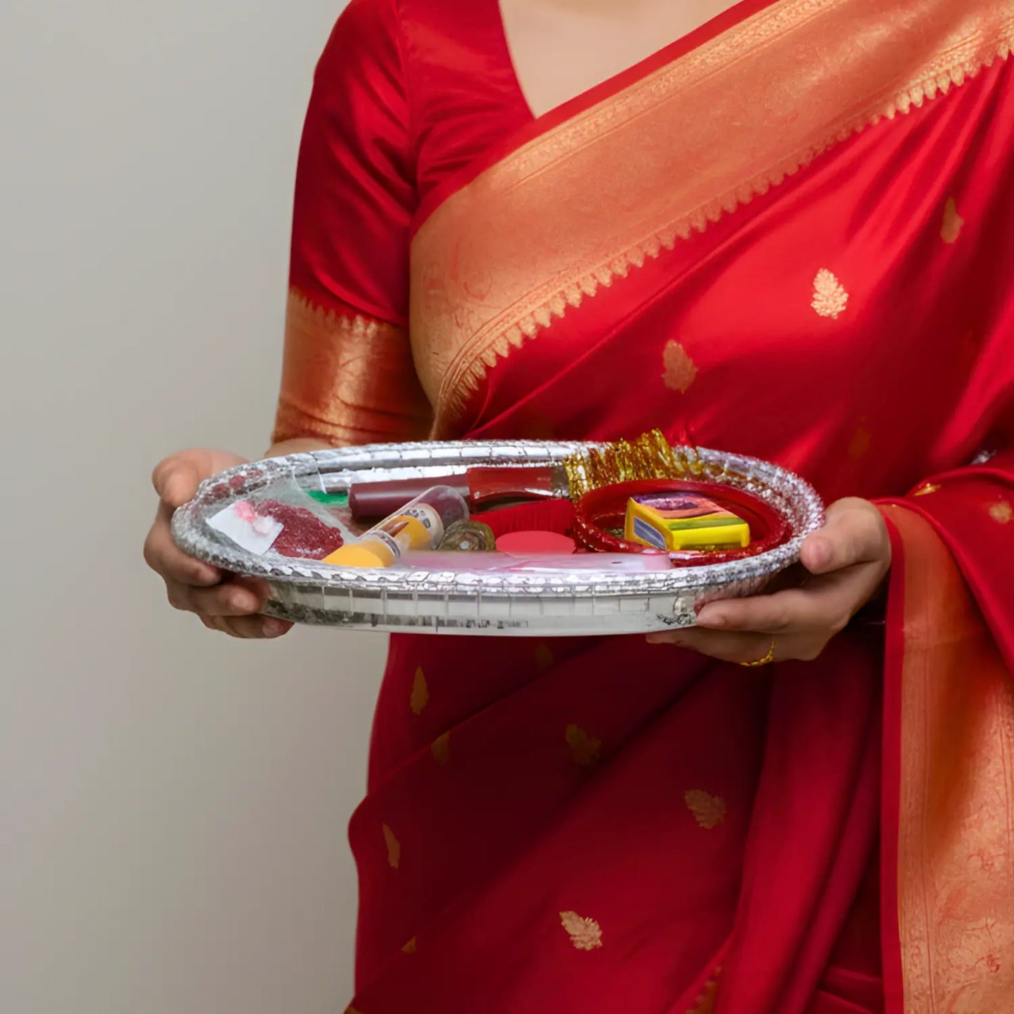 Person in a red saree holding a tray with various items against a plain background