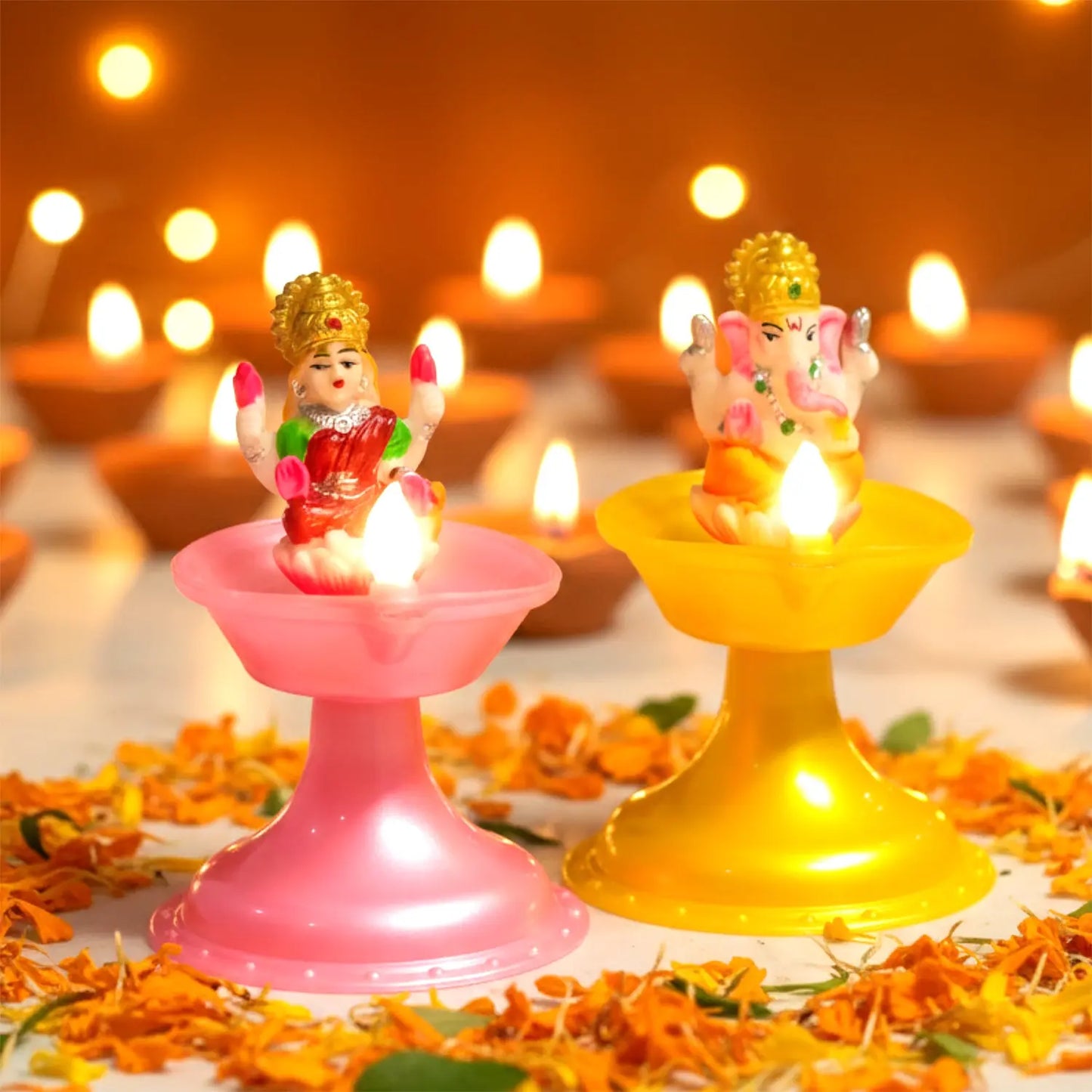Two small figurines on pink and yellow stands with candles, surrounded by flowers and blurred lights in the background. Water Sensor Laxmi ganesh Diya.