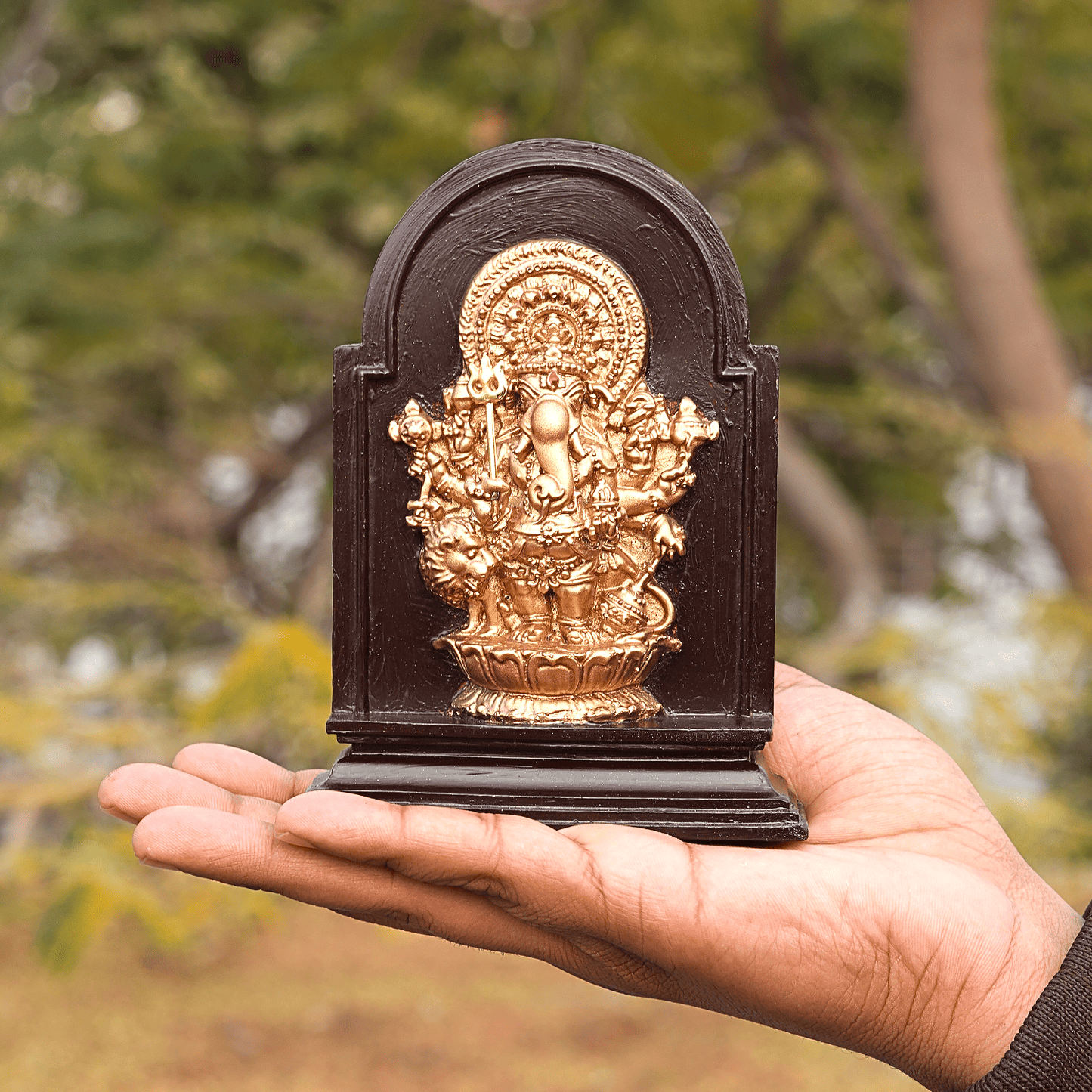 Hand holding a small golden deity statue on a dark wooden base against a blurred natural background
