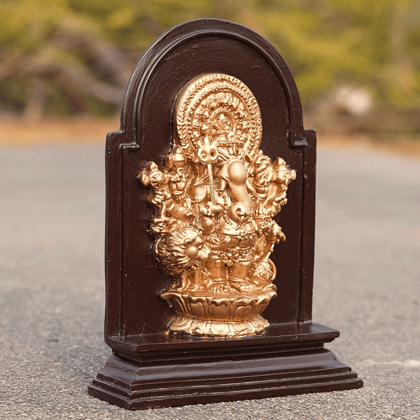 Gold-colored deity statue on a brown wooden base with a blurred natural background